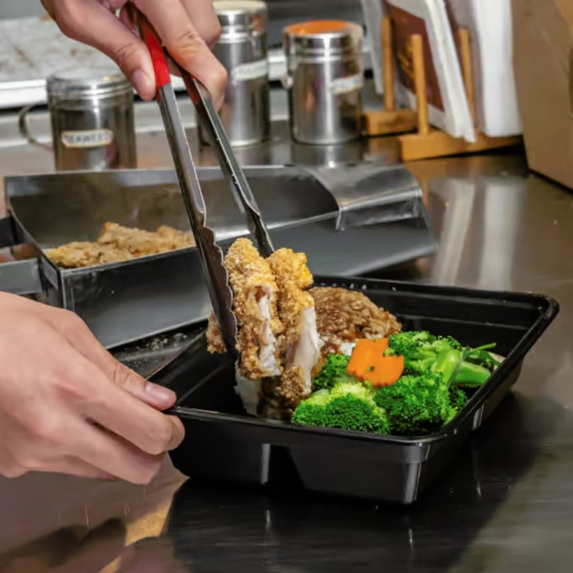 Hands using tongs to place crispy Taiwanese fried chicken into a takeout container with broccoli.